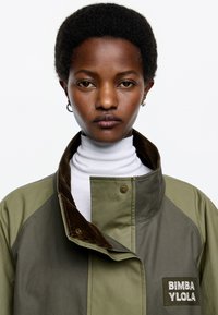 Chaqueta verde oliva con cuello alto, detalles de pana, cierres de presión metálicos y un parche con letras blancas que dice "BIMBA YLOLA."