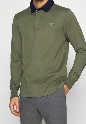 Mann trägt olivgrünes Langarm-Poloshirt mit marineblauem Kragen und Logo, kombiniert mit grauer Hose, steht vor weißem Hintergrund.