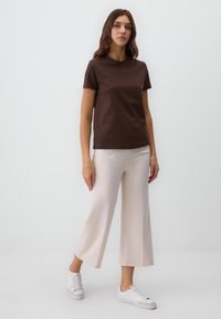 Braunes T-Shirt mit kurzen Ärmeln, kombiniert mit hellbeigen, weiten Hosen und weißen Turnschuhen. Der Stoff wirkt weich und hat ein schlichtes Design.