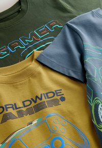 Drie katoenen t-shirts: donkergroen met de tekst "GAMER", mosterdgeel met "WORLDWIDE GAMER" en een ontwerp van een gamecontroller, en een blauwe graphic tee met een autoonwerp.