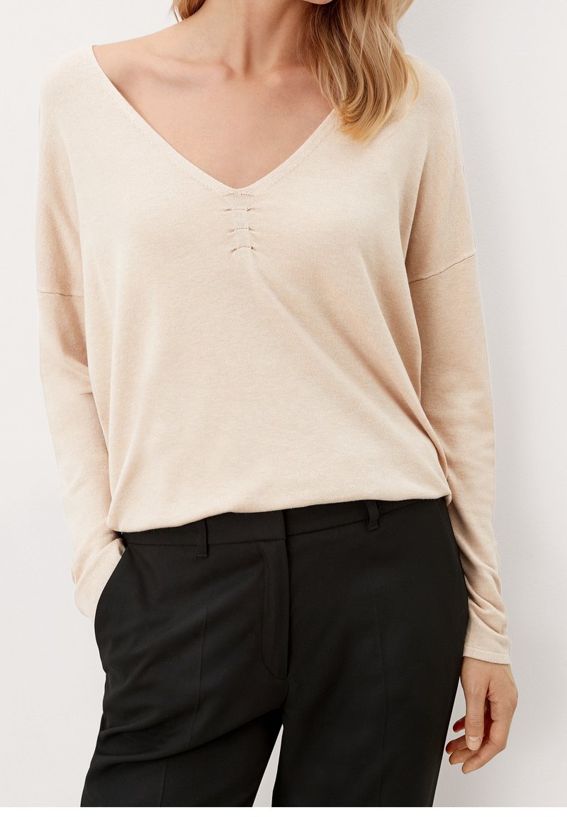 Beiger V-Ausschnitt-Pullover mit drei kleinen vertikalen Nähten in der Mitte, weicher Textur, langen Ärmeln, kombiniert mit schwarzen Hosen.