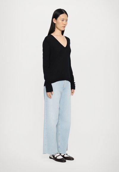 Zwarte V-halstrui met lange mouwen, gecombineerd met lichtblauwe wijde jeans. Model draagt zwarte schoenen met witte accenten. Eenvoudig, minimalistisch ontwerp.