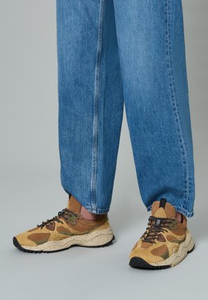Sneaker chunky marroni e beige con una finitura testurizzata, lacci neri e suole maculate, abbinati a pantaloni in denim blu larghi.