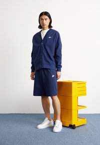 Temno modra Nike cardigan s gumbi, v kombinaciji z usklajenimi kratkimi hlačami. Model nosi bele športne copate in stoji ob rumeni enoti za shranjevanje.