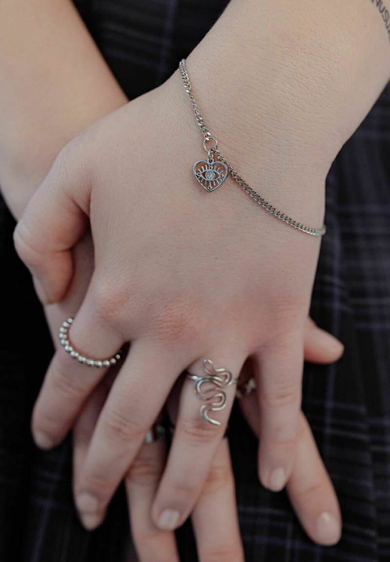 Silberarmband mit Herzanhänger an einer Hand, mit einem detaillierten Design. Zusätzliche silberne Ringe sind zu sehen, mit einer glatten Textur und einem glänzenden Finish.