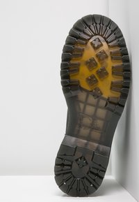 Semelle de botte en caoutchouc noire avec un motif de semelle épais, présentant une surface texturée et des formes géométriques pour l'adhérence. Comprend des détails de marque en relief.