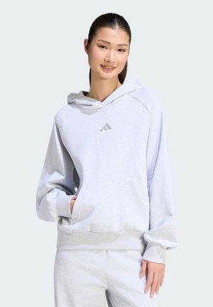 Jonge vrouw met donker haar die een lichtgrijze Adidas hoodie en bijpassende joggingbroek draagt, glimlachend met één hand in de zak.