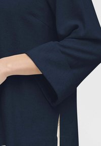 Top blu navy con finitura testurizzata, caratterizzato da maniche ampie e spaccature laterali, che mette in evidenza un design rilassato e una silhouette moderna.
