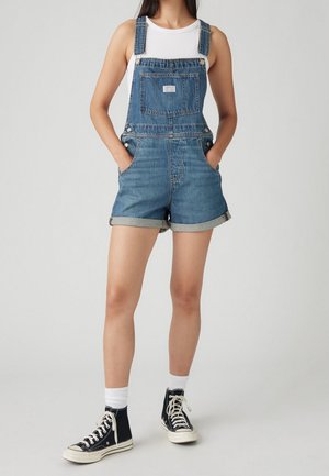 Femme portant une salopette courte en jean retroussée, un haut blanc sans manches, des chaussettes blanches et des baskets montantes noires, debout les mains dans les poches.