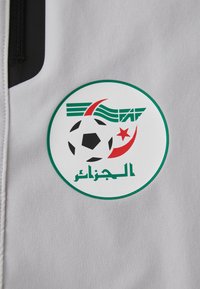 Κυκλείο λογότυπο που περιλαμβάνει μια ασπρόμαυρη μπάλα ποδοσφαίρου, με πράσινες και κόκκινες λεπτομέρειες, σε ανοιχτό γκρι υφασμάτινο φόντο. Περιλαμβάνεται αραβικό κείμενο.