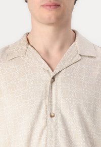 Camisa de manga corta beige con un sutil patrón de paisley blanco, que presenta un cuello con muesca y botones marrones en el delantero.