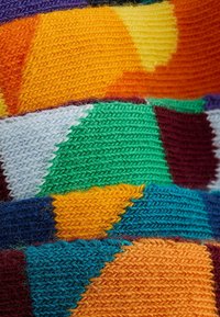 Tissu tricoté multicolore avec des motifs géométriques en orange, vert, bleu, violet et jaune. Mélange de laine texturé et doux avec des coutures visibles.
