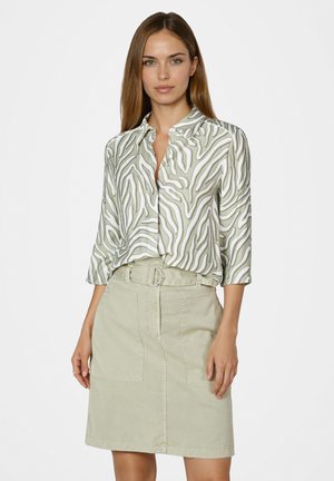 Frau trägt grün-weiß gemusterte Bluse mit Knöpfen und hellgrünen Rock mit Gürtel, steht vor einfarbigem Hintergrund.