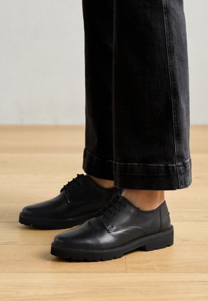 Czarne skórzane buty o gładkim wykończeniu, z zaokrąglonym noskiem, sznurowane, z teksturowaną gumową podeszwą, noszone z ciemnymi dżinsami.