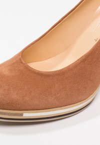 Escarpin en suède marron à bout arrondi, avec une semelle extérieure striée en couches beige, marron et blanche. Doublure intérieure lisse.