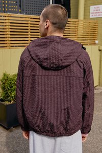 Giacca a quadri bordeaux con cappuccio, tessuto testurizzato, orlo elastico e rifiniture a contrasto. Il modello indossa pantaloni corti grigi; piante verdi sullo sfondo.