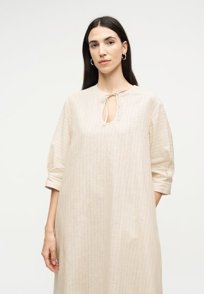 Γυναίκα με μακριά σκούρα μαλλιά, φορώντας ένα άνετο μπεζ φόρεμα με λεπτές κάθετες ρίγες και κλείσιμο με κορδόνι στο ντεκολτέ, στέκεται μπροστά σε λευκό φόντο.