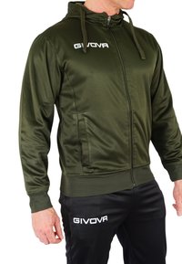 Giacca verde con zip realizzata in tessuto lucido, dotata di cappuccio, tasche laterali e polsini a coste, abbinata a pantaloni neri con logo bianco.