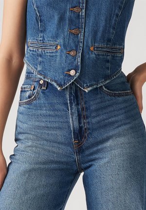Close-up of a person wearing a buttoned blue denim vest and high-waisted blue denim jeans with a hand resting on the hip.