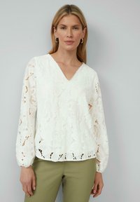 Blouse en dentelle blanche avec un col en V, des manches longues bouffantes, des motifs floraux et un ourlet festonné. Associée à un pantalon vert olive.
