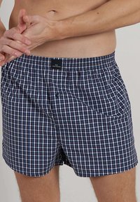 Checkered boxers in navy, white, and burgundy fabric. Features an elastic waistband, button closure, and side pockets for storage.