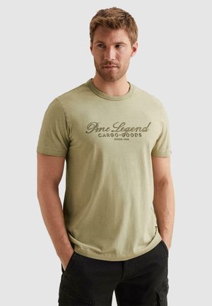 Man with short hair wearing a light olive green "Pme Legend Cargo Goods" T-shirt and black cargo pants, hands in pockets.