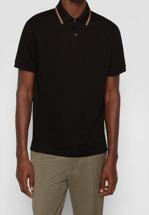 Mężczyzna w czarnej polo z krótkim rękawem, z wielokolorowym paskowanym kołnierzem i beżowymi spodniami, stojący na jednolitym tle.