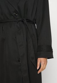 Trench-coat noir avec un design structuré, fermeture à boutons, ceinture à la taille et manches légèrement fuselées avec des détails accentués.