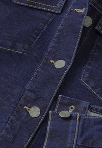 Mörkblå denimtyg med kontrasterande gula sömmar, utrustad med knäppningar och en design med bröstficka. Slät textur och robust konstruktion.