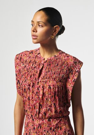 Frau mit hochgestecktem Haar, die eine rosa gemusterte ärmellose Bluse mit Falten und kleinen Knöpfen trägt, schaut nach links vor einfachem Hintergrund.