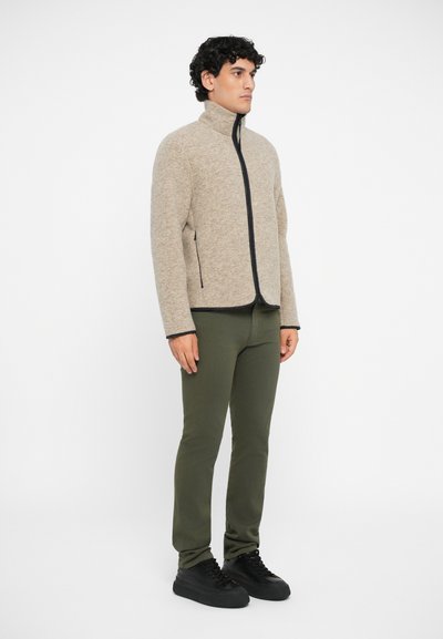 Beige zip-up fleece jacket with a high collar, black zipper, and side pockets. Pair of slim-fit olive green pants and black sneakers.