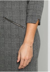 Robe à carreaux noir et blanc avec un détail de poche zippée, manches courtes et tissu texturé. Bracelet argenté délicat au poignet.