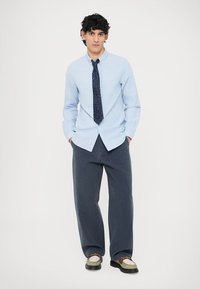 Φωτεινό γαλάζιο πουκάμισο με κουμπιά και τυπικό γιακά, συνδυασμένο με σκούρα παντελόνια φαρδιάς γραμμής. Το σύνολο περιλαμβάνει μια ναυτική γραβάτα με πουά μοτίβο και παπούτσια με δίχρωμο σχέδιο.