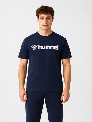 Młody mężczyzna stoi, ubrany w granatowy t-shirt z białym logo "hummel" i dopasowane granatowe spodnie na jednolitym jasnym tle.