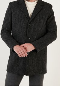 Abrigo negro texturizado con un diseño entallado, cierre de un solo botón y dos botones. Presenta un cuello de muesca y bolsillos laterales.