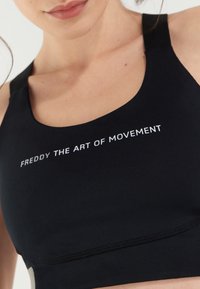 Melna sporta krūšturis, kas izgatavots no elastīga auduma, ar apaļu kakla izgriezumu un tekstu "FREDDY THE ART OF MOVEMENT" baltām burtiem priekšpusē.