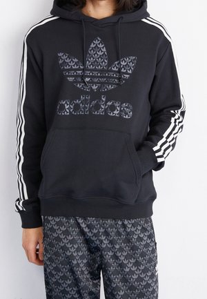 Person trägt schwarzen Adidas Hoodie mit weißen Streifen an den Ärmeln und passenden gemusterten Hosen mit Adidas-Logos.