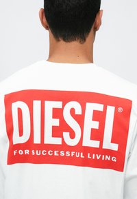 Sweatshirt branco com grande texto gráfico vermelho "DIESEL" e texto menor "PARA UMA VIDA SUCESSFUL" nas costas. Mangas curtas, ajuste casual.
