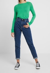 Maglia verde a maniche lunghe con texture a coste, abbinata a jeans blu a vita alta, con orli arrotolati, e scarpe da ginnastica nere con suole bianche.