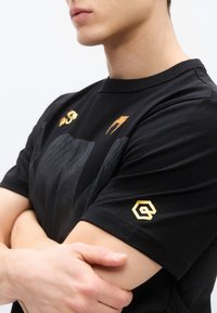Camiseta de algodón negra con mangas cortas, con logotipos dorados en el pecho y la manga izquierda, y un patrón texturizado en la parte frontal.