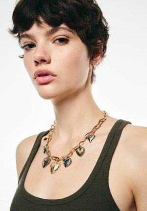 Jeune personne avec les cheveux bouclés courts portant un collier en chaîne dorée épaisse avec des pendentifs en forme de cœur et un débardeur vert olive.