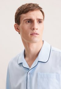 Chemise de pyjama bleu clair avec un col pointu, un liseré navy, une poche poitrine et une fermeture à boutons sur la patte de boutonnage. Texture de tissu douce.