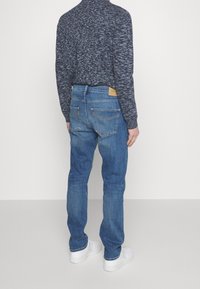 Personne debout de dos, portant un jean bleu, un pull bleu foncé à motifs et des baskets blanches sur un fond neutre.