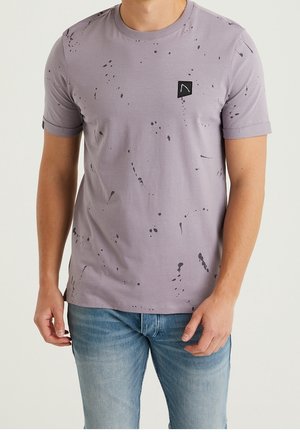 Mann trägt ein hellviolettes kurzärmeliges T-Shirt mit schwarzem Farbspritz-Design und kleinem schwarzem Aufnäher, kombiniert mit hellblauen Jeans.