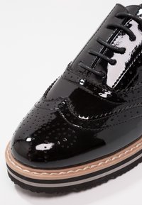 Chaussure en cuir verni noir avec un design à bouts rapportés, comportant des perforations et une semelle superposée avec des rayures beige et noires. Fermeture à lacets.