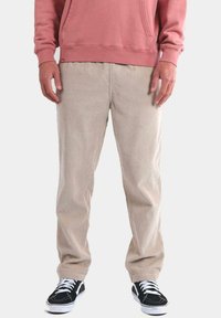 Pantalones de pana beige con un diseño de pierna recta, que presentan un ribete vertical y un ajuste relajado, combinados con zapatillas negras y una sudadera rosa.