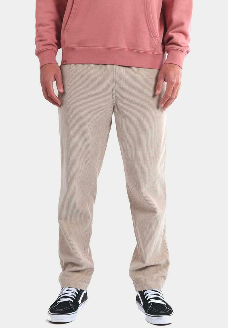 Pantalones de pana beige con un diseño de pierna recta, que presentan un ribete vertical y un ajuste relajado, combinados con zapatillas negras y una sudadera rosa.