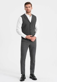 Grijze, gestructureerde vest over een wit shirt, gecombineerd met slim-fit grijze broeken en zwarte schoenen. Knopen en zakken sieren het ontwerp van de vest.