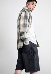 Plaid shirt in beige and blue tones layered over a white t-shirt, paired with black denim shorts featuring multiple pockets and branded patch.