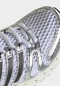 Chaussure de sport blanche avec des superpositions argentées, des lacets blancs et une tige en mesh respirant à motif alvéolé.